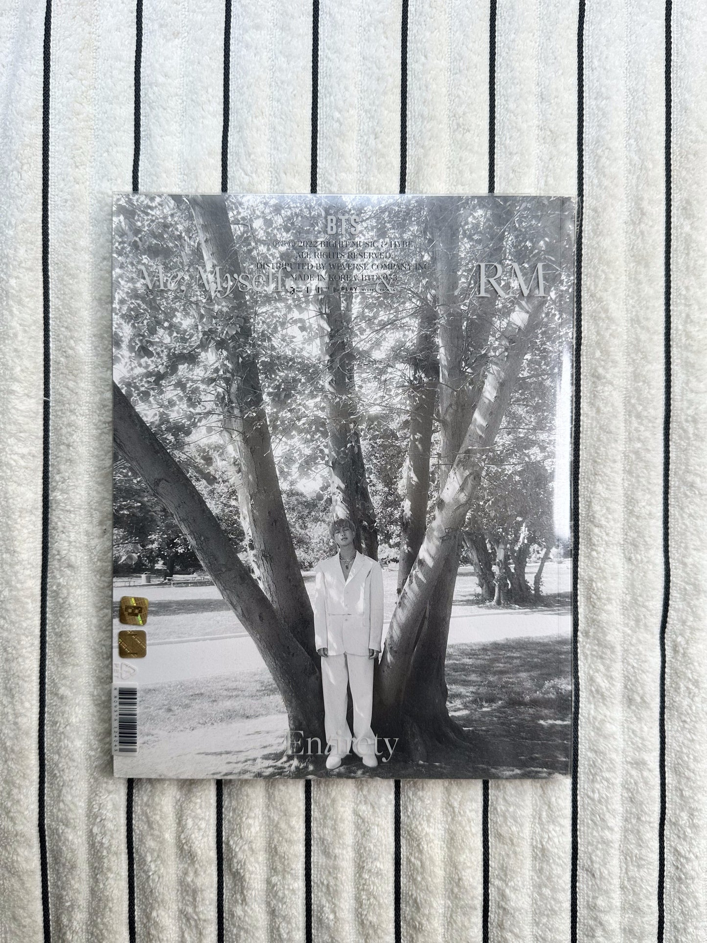 A rectangular printed magazine or book cover laid flat on a textured white fabric background with thin dark horizontal lines. The cover is mostly black-and-white photography of tree trunks and branches, with small gold logo blocks near the bottom right.