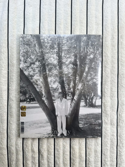 A rectangular printed magazine or book cover laid flat on a textured white fabric background with thin dark horizontal lines. The cover is mostly black-and-white photography of tree trunks and branches, with small gold logo blocks near the bottom right.