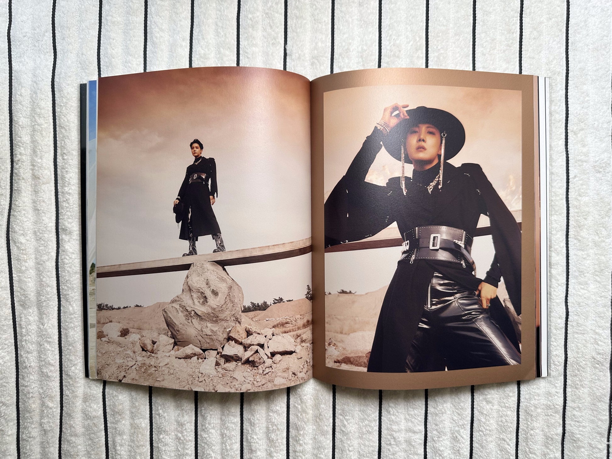 An open magazine or photo book laid flat on a white textured fabric background, showing two printed fashion photographs side by side. The top page features a close-up of a person in a black outfit and hat, and the bottom page shows a full-body shot of the same person standing outdoors near rocks.