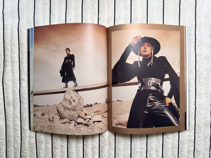 An open magazine or photo book laid flat on a white textured fabric background, showing two printed fashion photographs side by side. The top page features a close-up of a person in a black outfit and hat, and the bottom page shows a full-body shot of the same person standing outdoors near rocks.