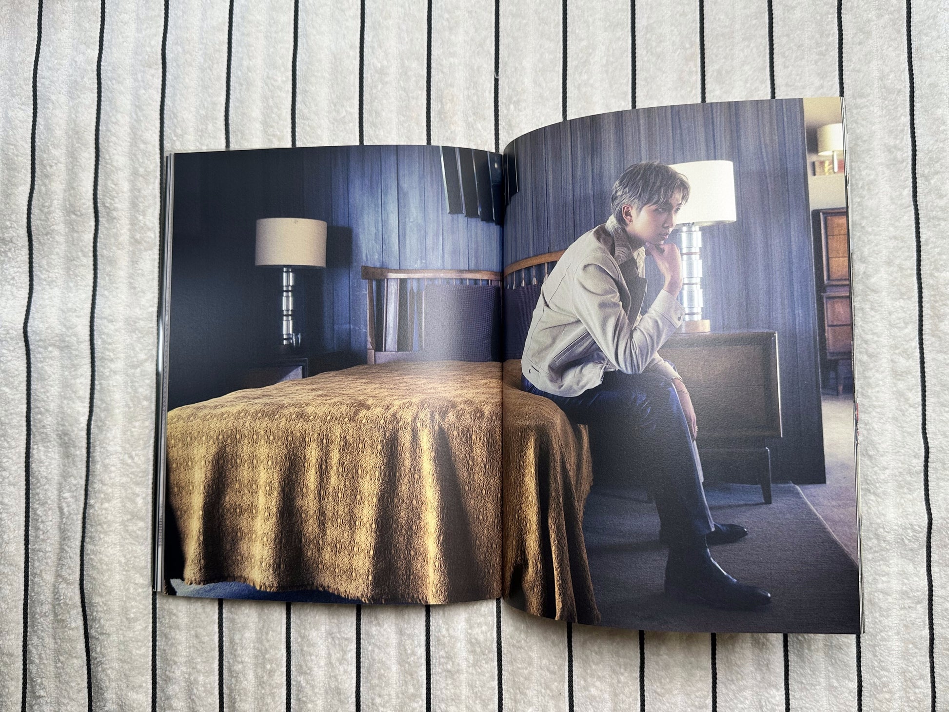 An open magazine or photo book lying on a gray striped fabric surface, showing a printed interior scene with a person in a room and a large gold draped textile.