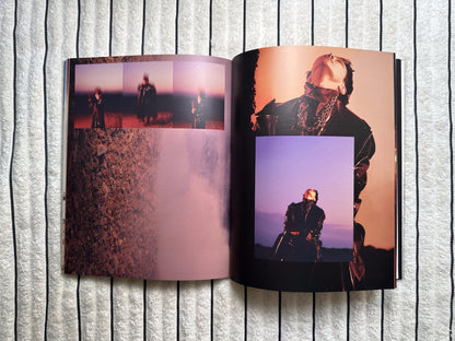 An open photo book or printed album spread laid flat on a light gray ribbed surface. The pages contain multiple artistic photographs with warm and cool tones, including a figure in dark clothing against an orange sky and a purple-toned portrait.