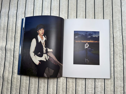 An open printed photo album or photo book laid flat on a gray textured fabric background. The top page shows a small square black-and-white scenic photo, and the open lower page shows a larger portrait-style photo of a person in a dark outfit.