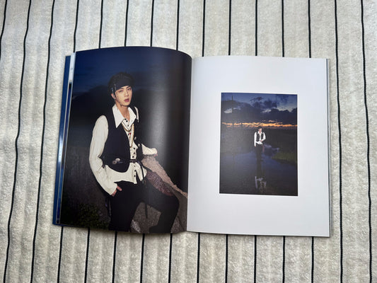 An open printed photo album or photo book laid flat on a gray textured fabric background. The top page shows a small square black-and-white scenic photo, and the open lower page shows a larger portrait-style photo of a person in a dark outfit.